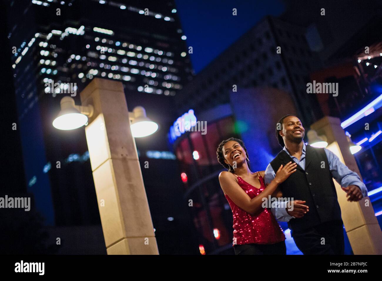 Happy young couple talking and laughing as they walk arm in arm through the city at night. Stock Photo