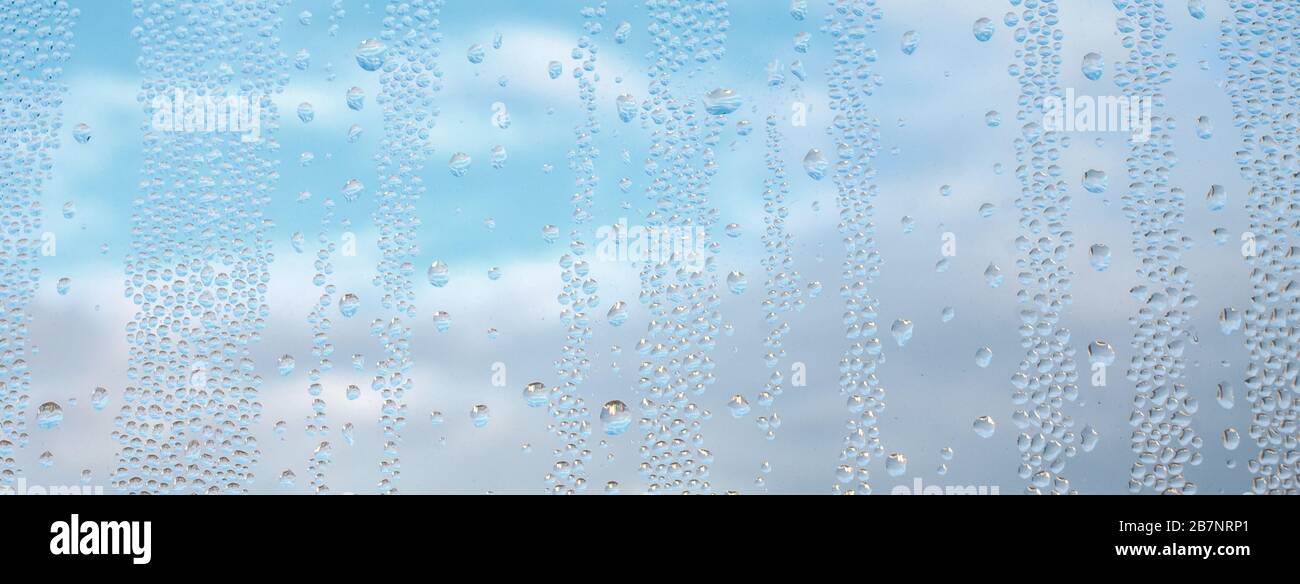 Drops water on window for wallpaper design. Blue cloudy sky backdrop. Spotted abstract texture background. Spring wet weather panorama. Droplets on glass in horizontal natural cover. Fit to autumn. Stock Photo