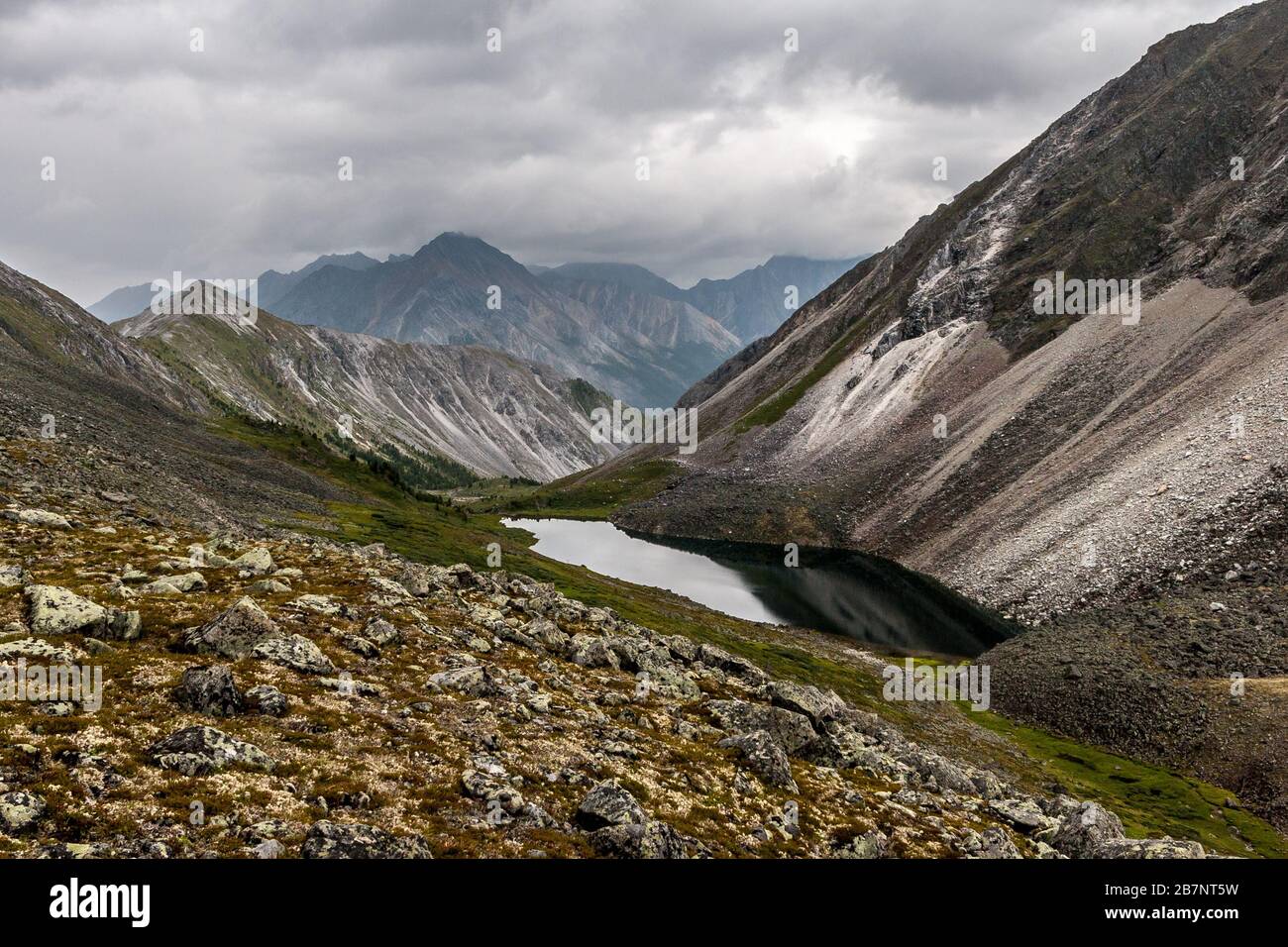 Mountain lake with a rocky slope in the foreground and mountain ranges in the background. Moss on the stones. Clouds in the sky. Horizontal. Stock Photo