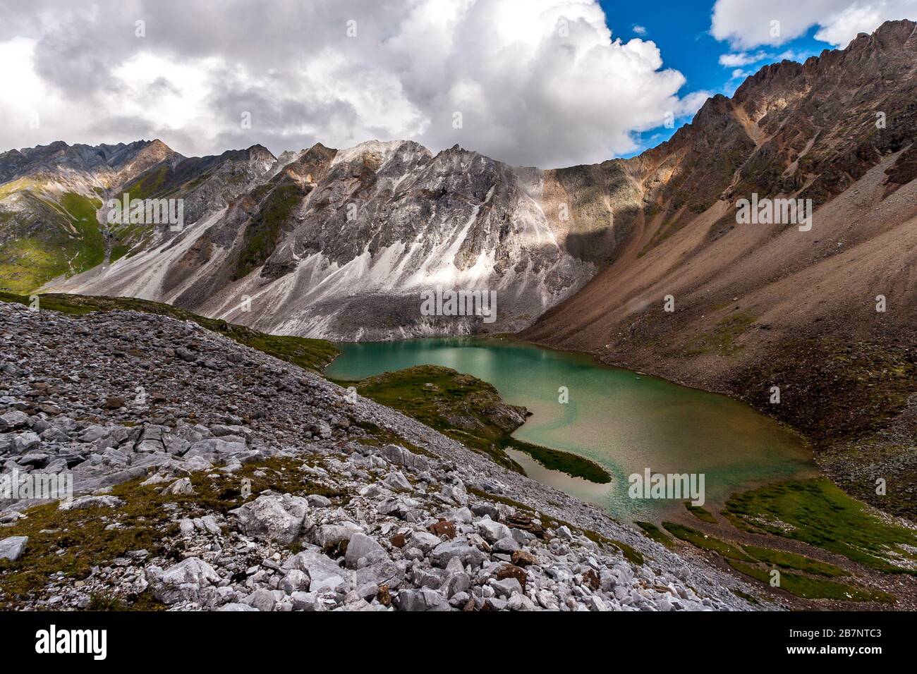 A small lake among the mountains. White and brown mountain nearby. Stone scree in the foreground. White clouds on a blue sky. Horizontal. Stock Photo