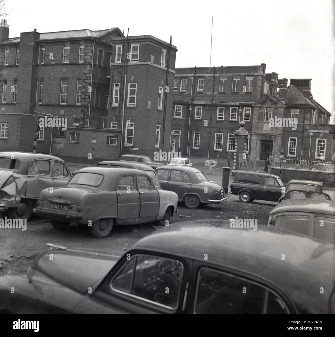 1960s, historical, cars of the era, many of them damaged and abandoned, parked on rough or waste ground, outside a victorian hospital building, South East London, England, UK. Stock Photo