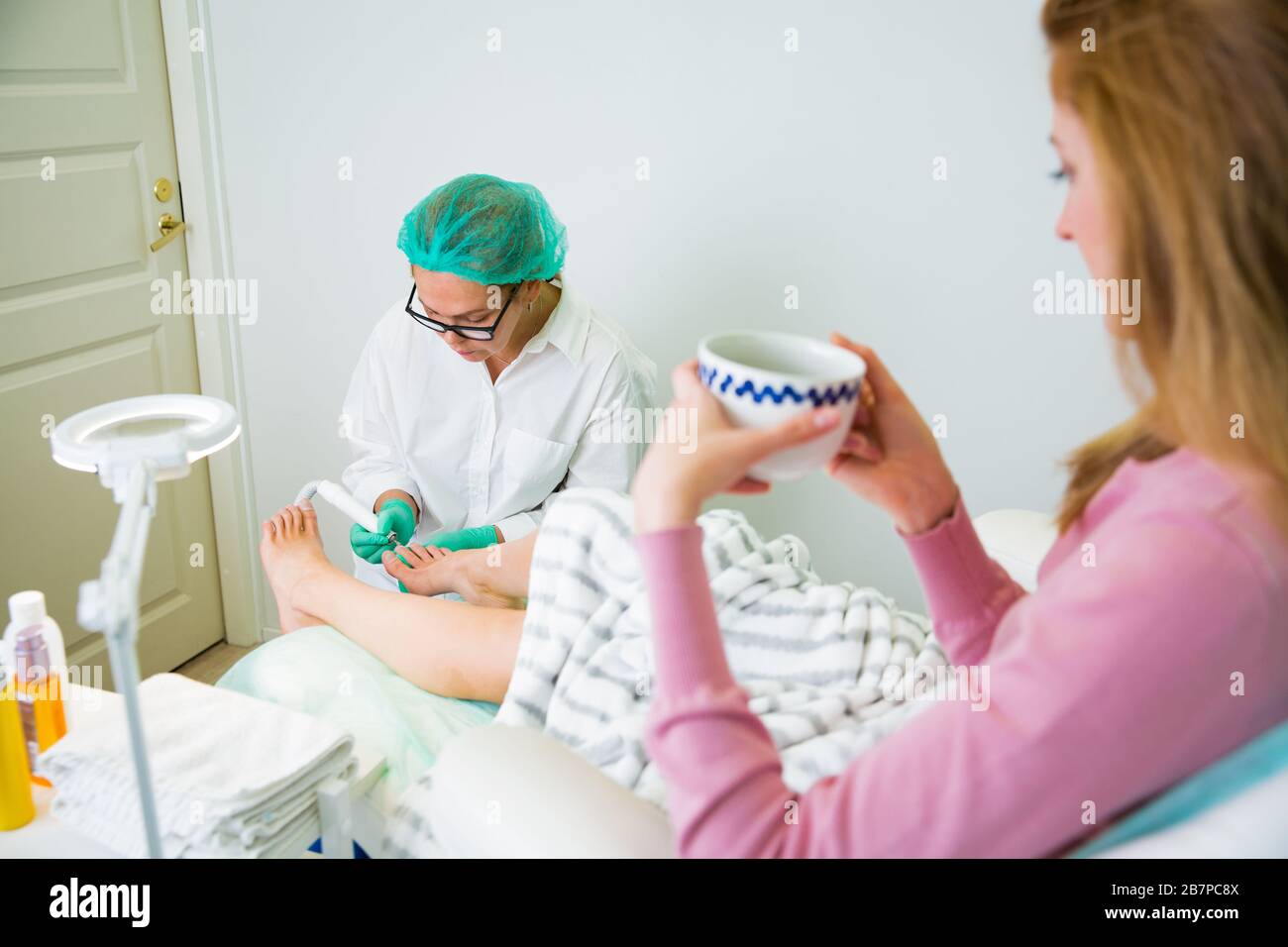 Woman relaxing in chair with cup of coffee, having foot treatment in spa salon.  Professional medical pedicure specialist in protective mask Stock Photo
