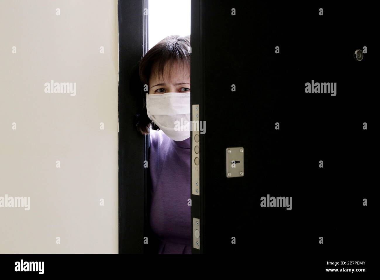 Home quarantine during the COVID-19 coronavirus epidemic. Worried woman in a medical mask standing in the open doorway Stock Photo