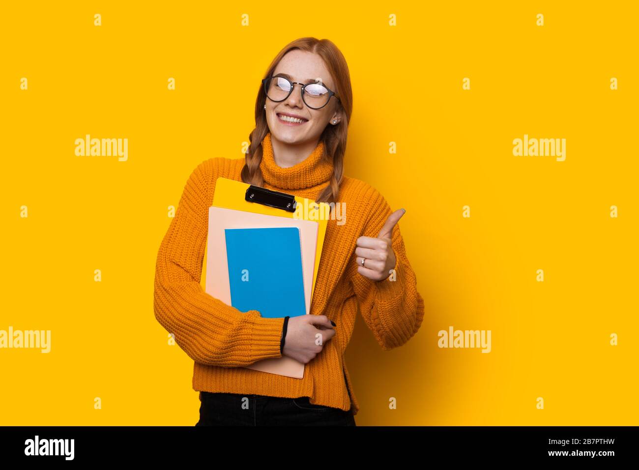 Happy red haired caucasian student with freckles and eyeglasses is holding some books and gesturing the like sign on a yellow background Stock Photo