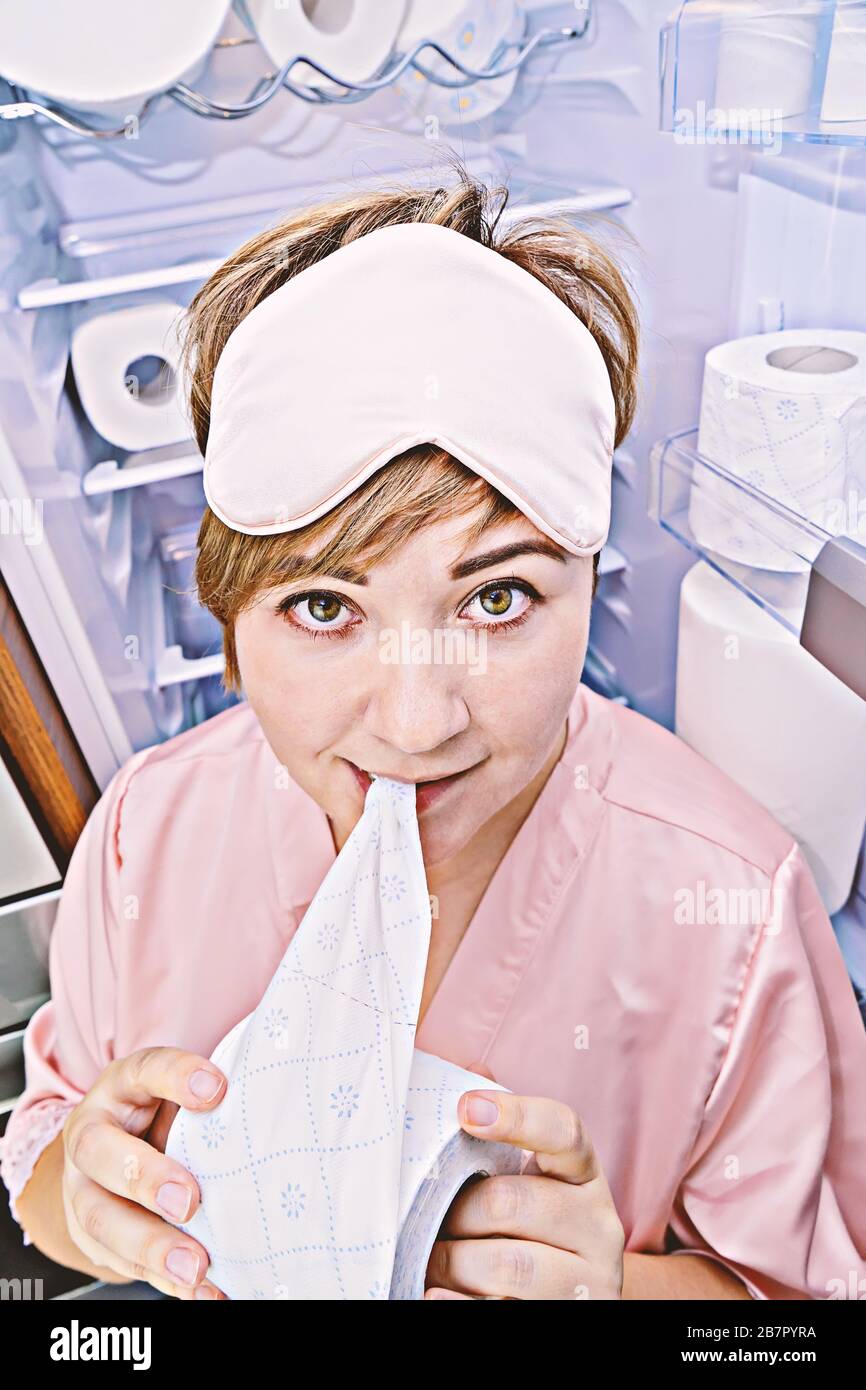 Woman in pajamas eagerly eats toilet paper near the fridge filled with