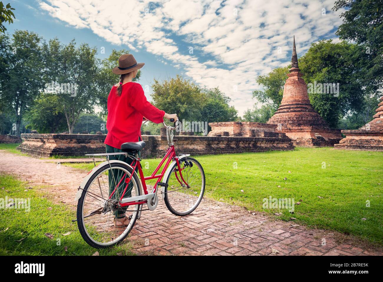 Woman in red shirt with vintage bicycle looking at old ruined Buddhist temple in amazing Sukhothai historical park, Thailand Stock Photo
