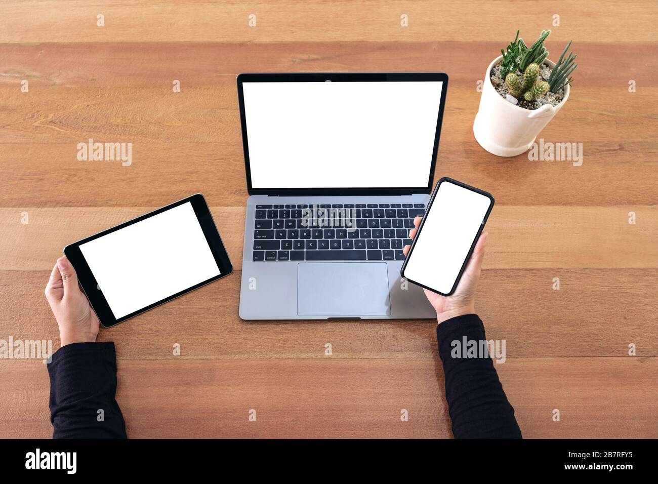 Top view mockup image of a hands holding mobile phone , black tablet and laptop with blank white screen on wooden table in office Stock Photo