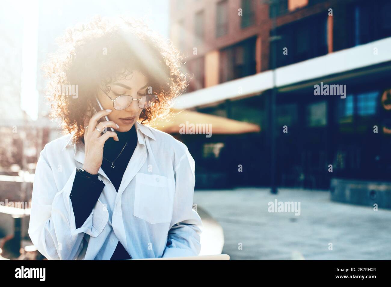 Stunning caucasian woman with curly hair and eyeglasses is wearing a white shirt and having a phone discussion Stock Photo