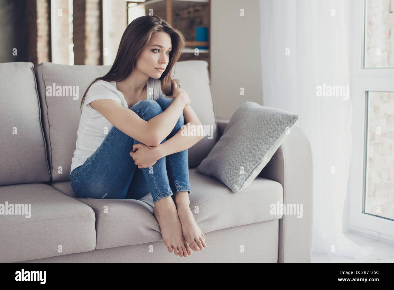 Young pretty lonely sad girl sitting on sofa and hugging her knees ...