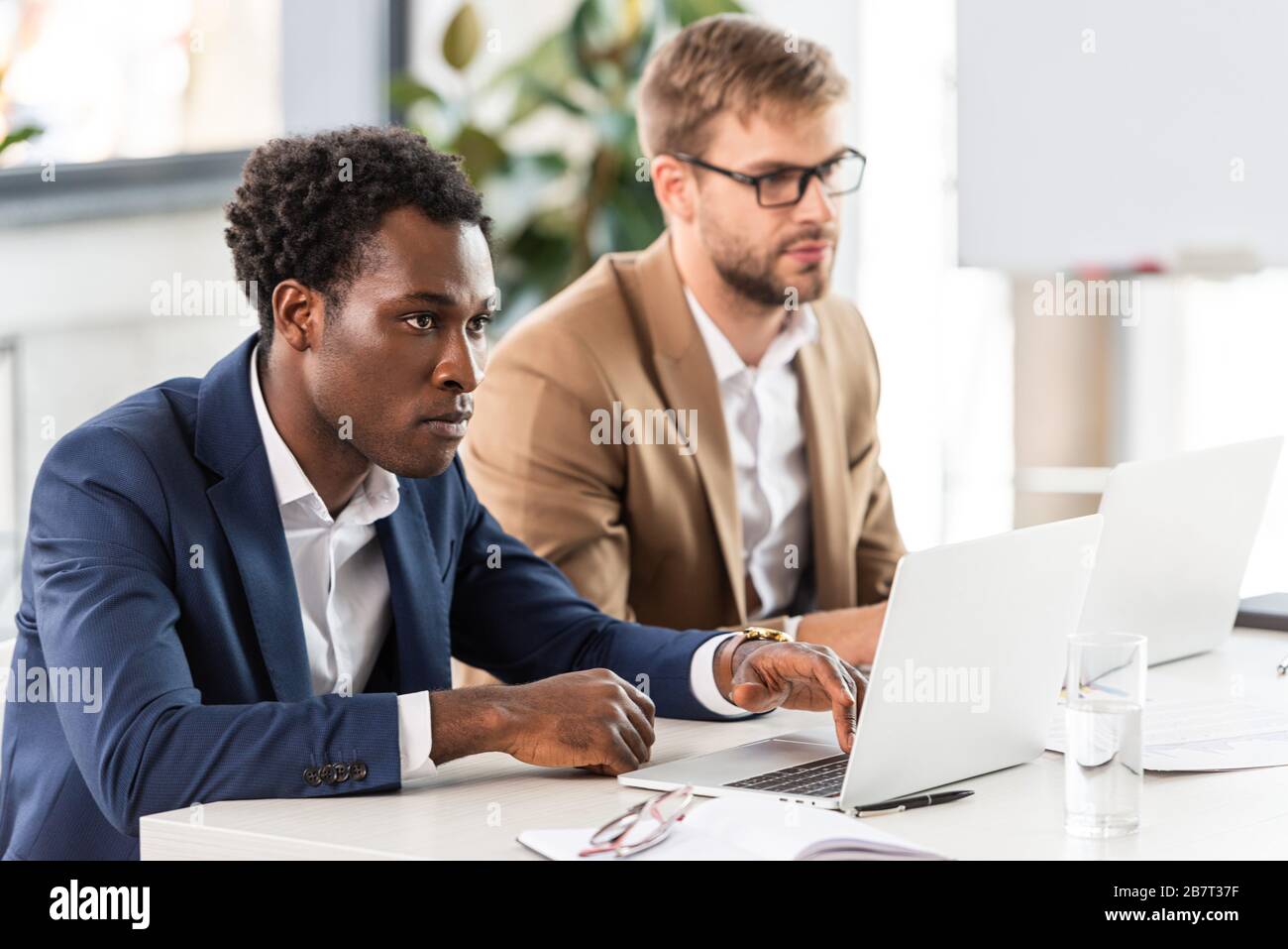 two multiethnic businessmen using laptops at table in office Stock Photo