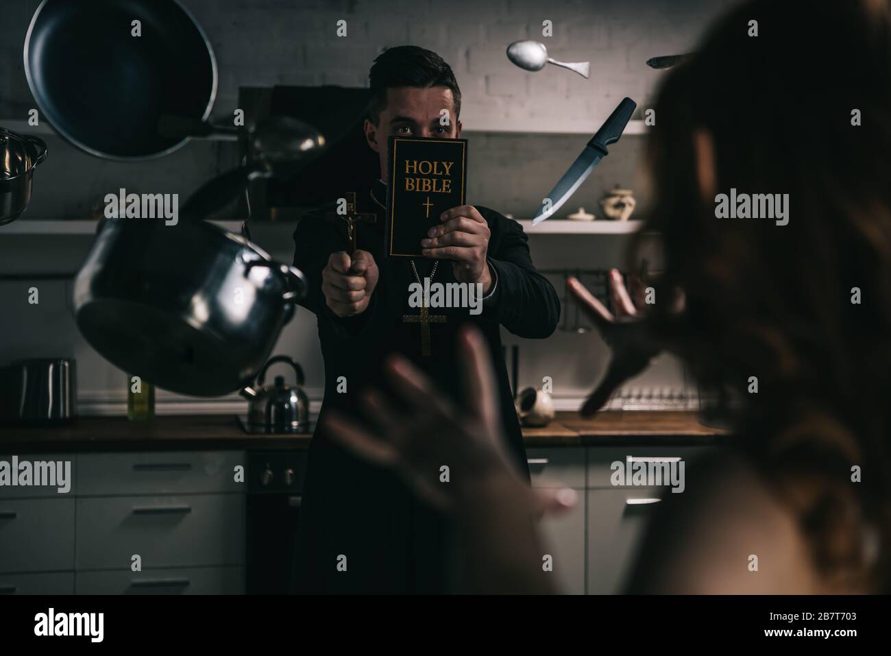 selective focus of demon with levitating cookware and exorcist with cross and bible in kitchen Stock Photo