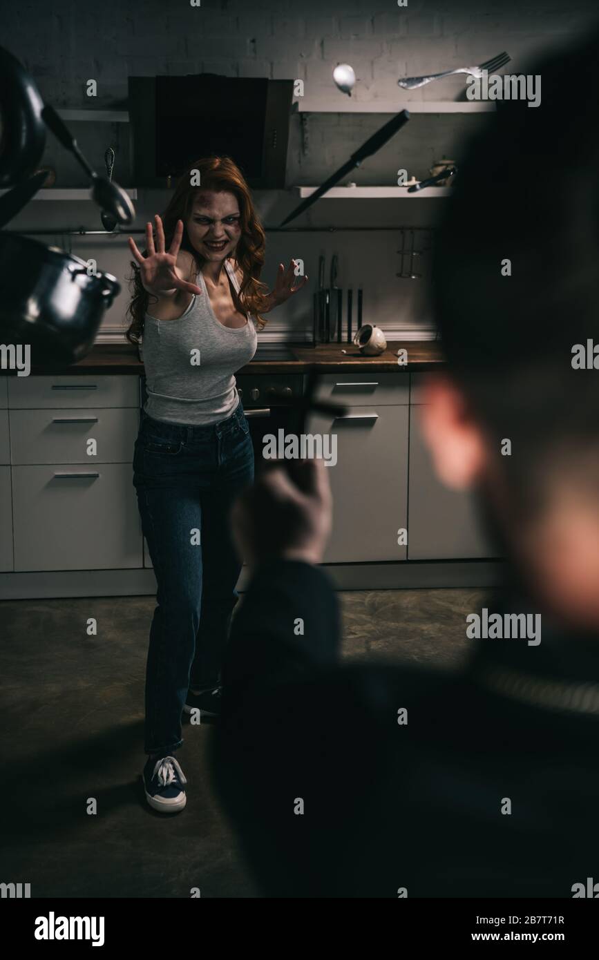 selective focus of creepy demoniacal girl with levitating cookware and exorcist with cross in kitchen Stock Photo