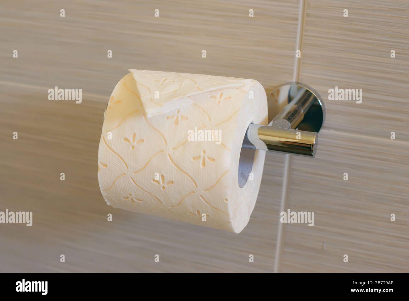 Toilet paper roll with over orientation and fold, set on a metallic holder or a bathroom tissue dispenser against a neutral tiled wall Stock Photo
