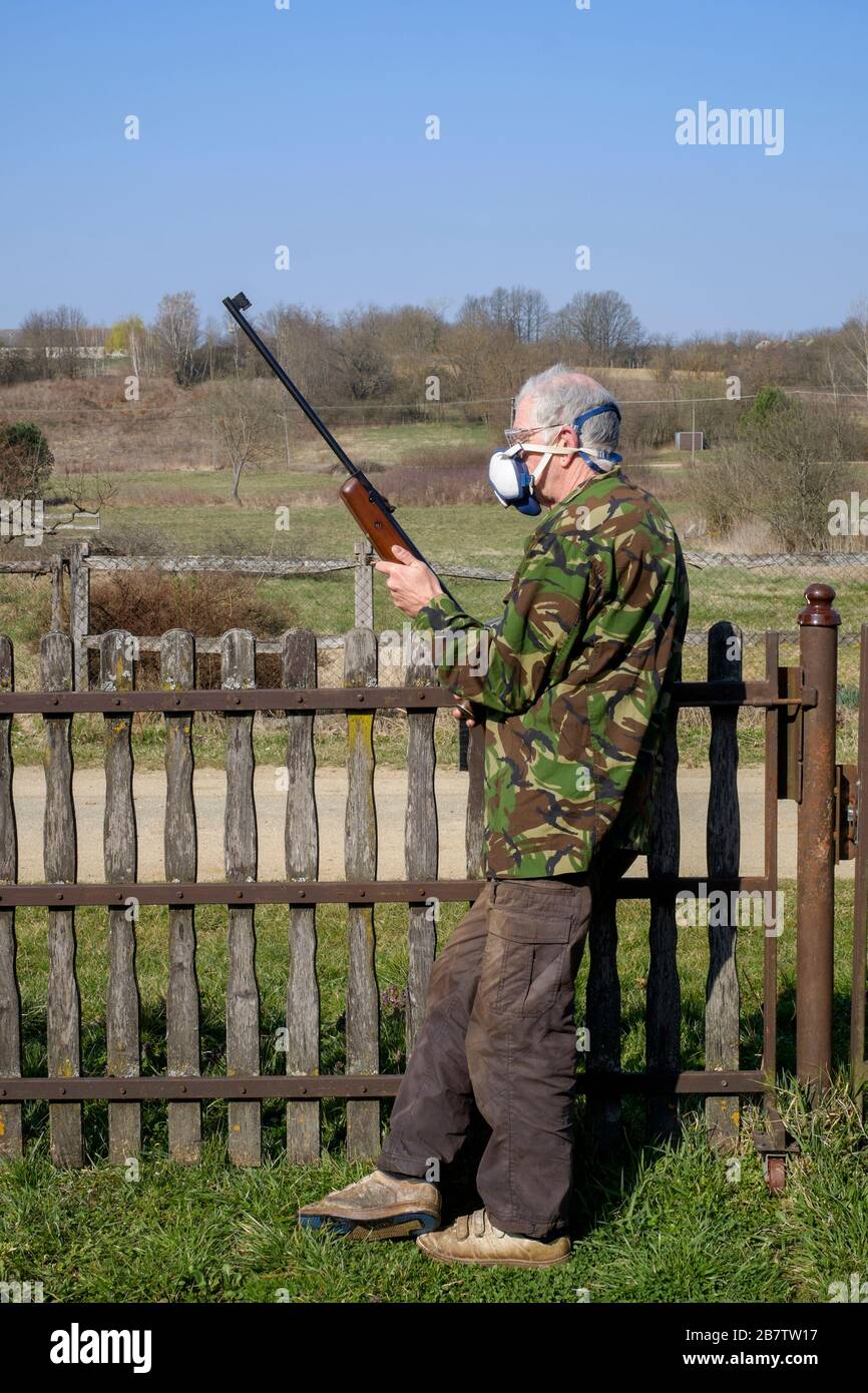 man wearing a face mask as protection against coronavirus infection standing with a rifle by garden gate in an isolated rural location hungary Stock Photo
