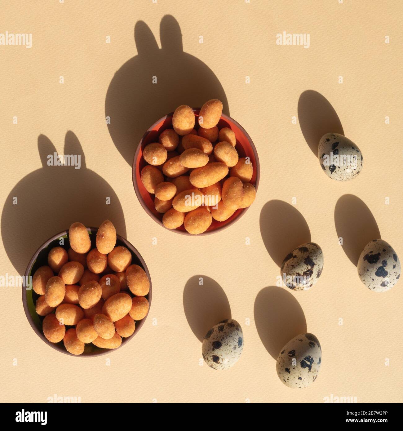 Five quail eggs and two bowls with nuts with shadows like rabbits on the light yellow background. Trendy conceptual minimalistic photo for Easter Holi Stock Photo