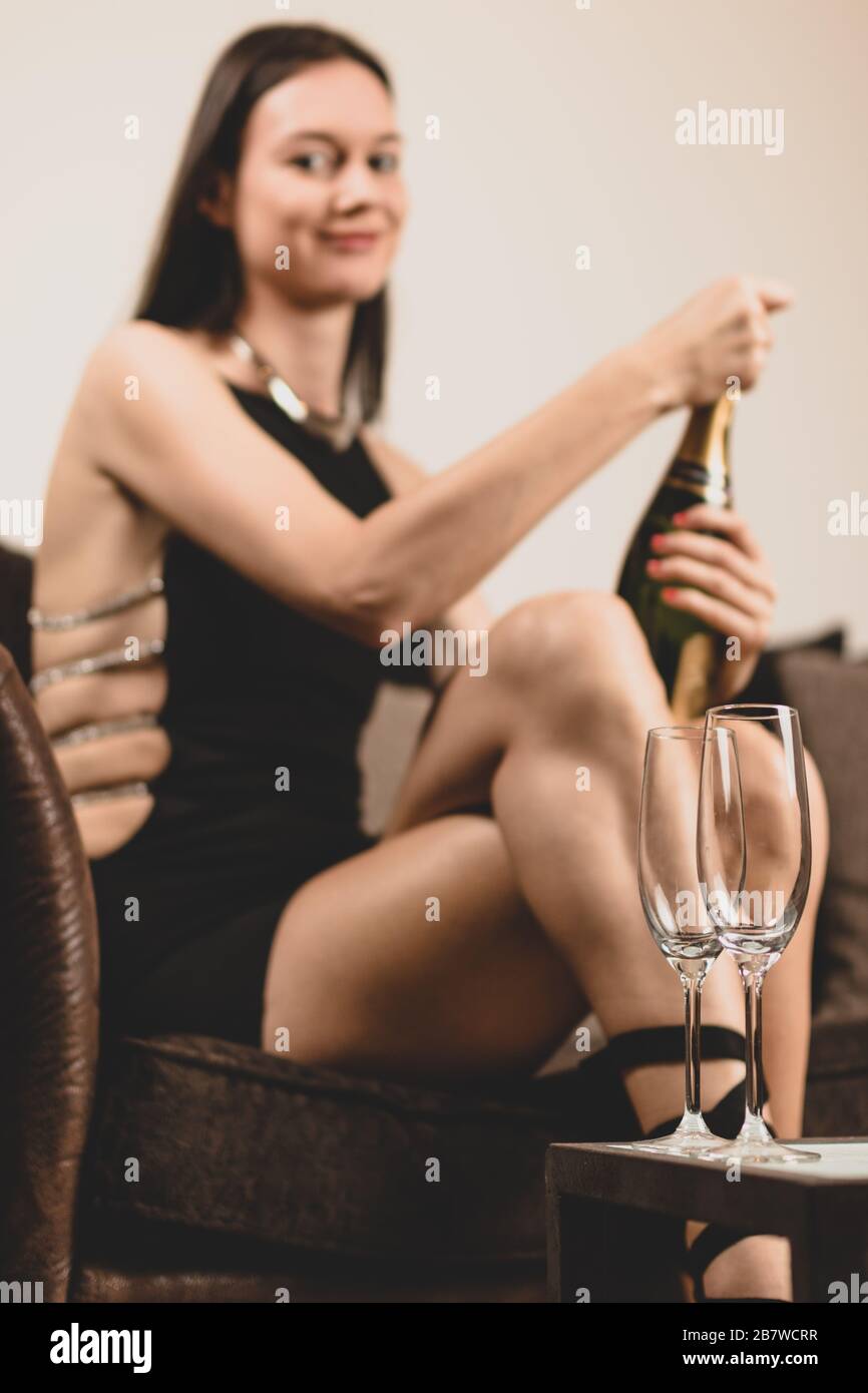 Classy young woman wearing a fancy dress, sitting on couch in a lounge, opening a champagne bottle. Royalty free stock photo. Stock Photo