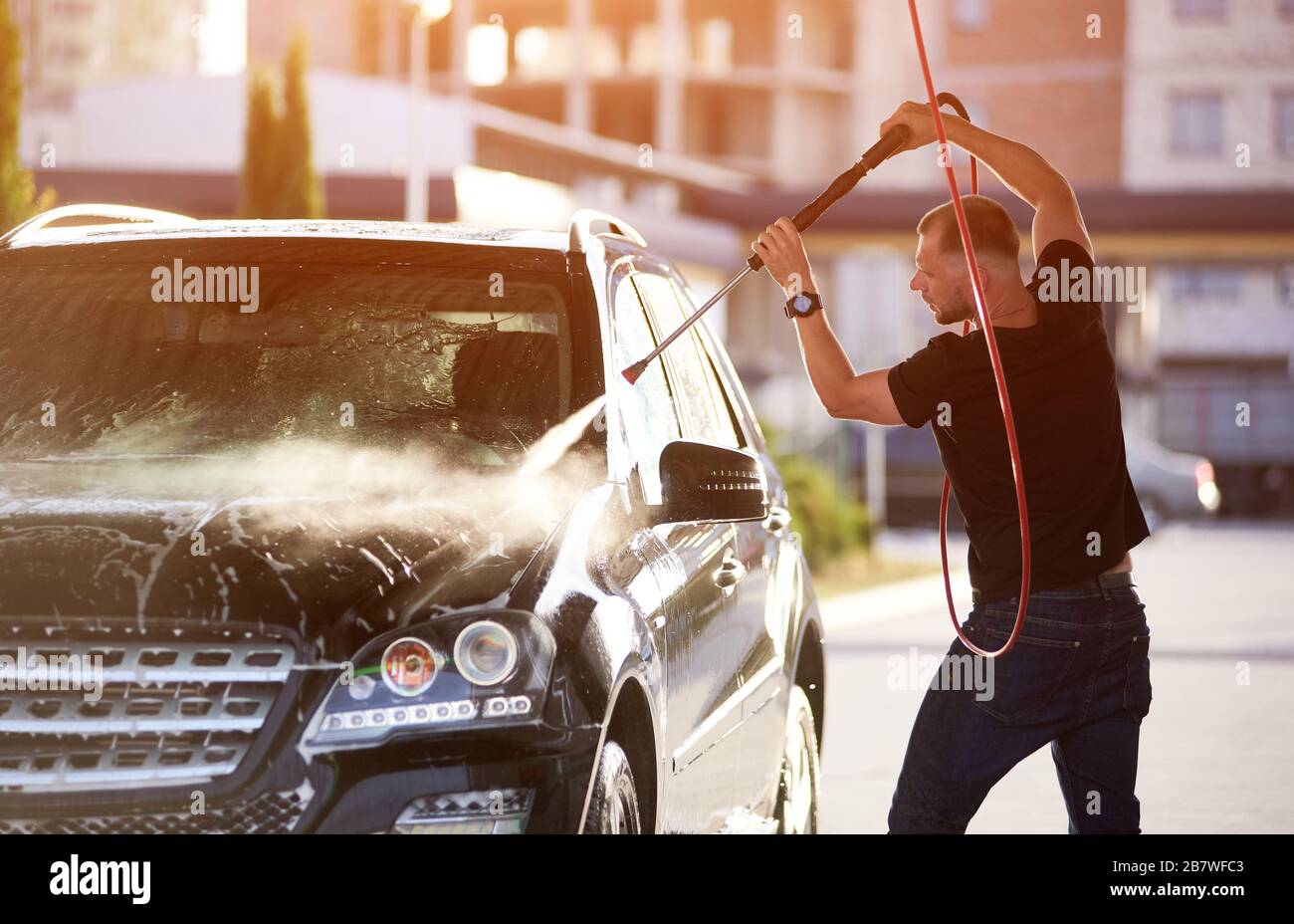 Side view of a man washing his car, holding a hose over his head, water jet under high pressure cleaning black car, surface of the vehicle is shining on the sun Stock Photo