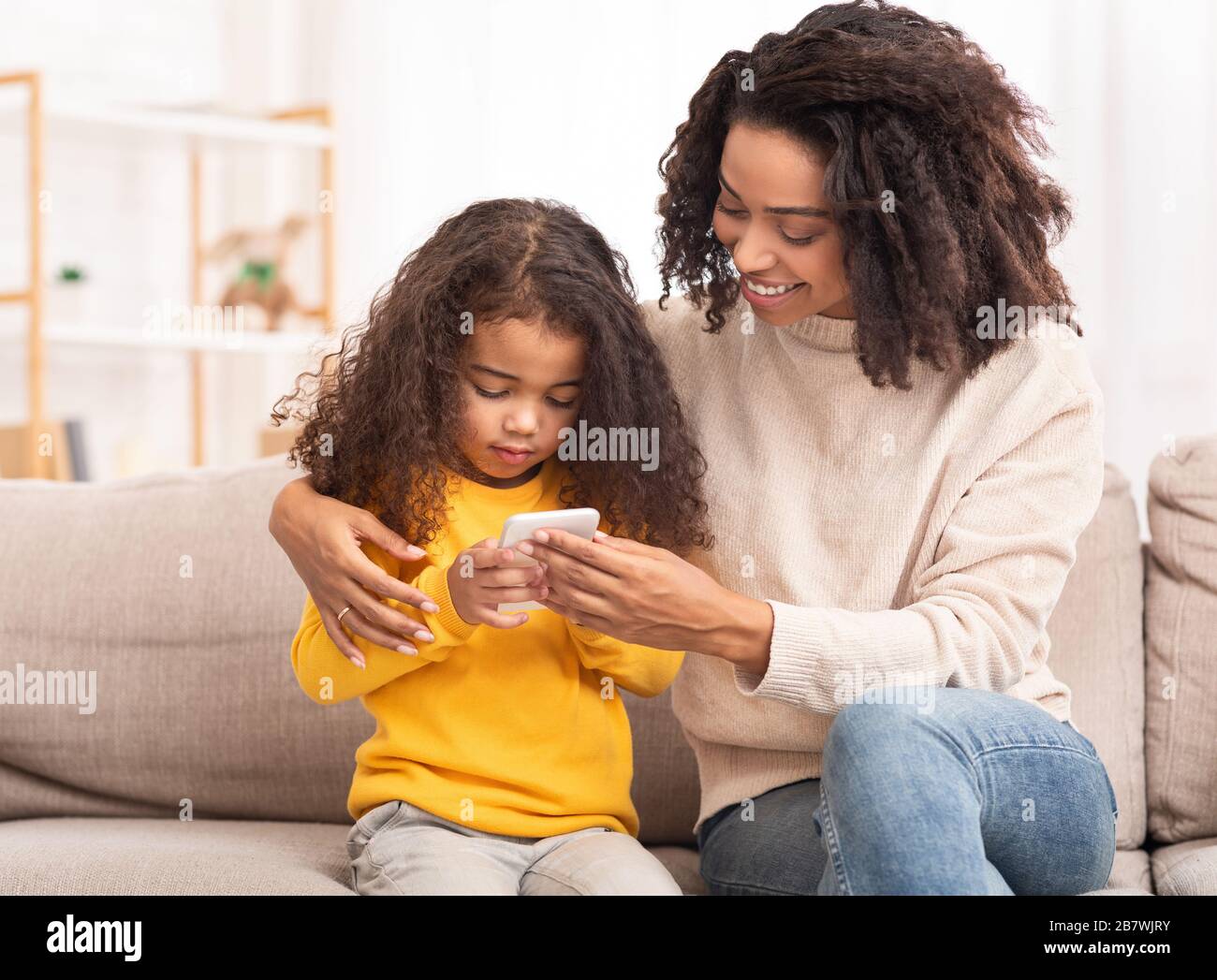 African Mother Teaching Daughter To Use Smartphone Sitting On Sofa Stock Photo