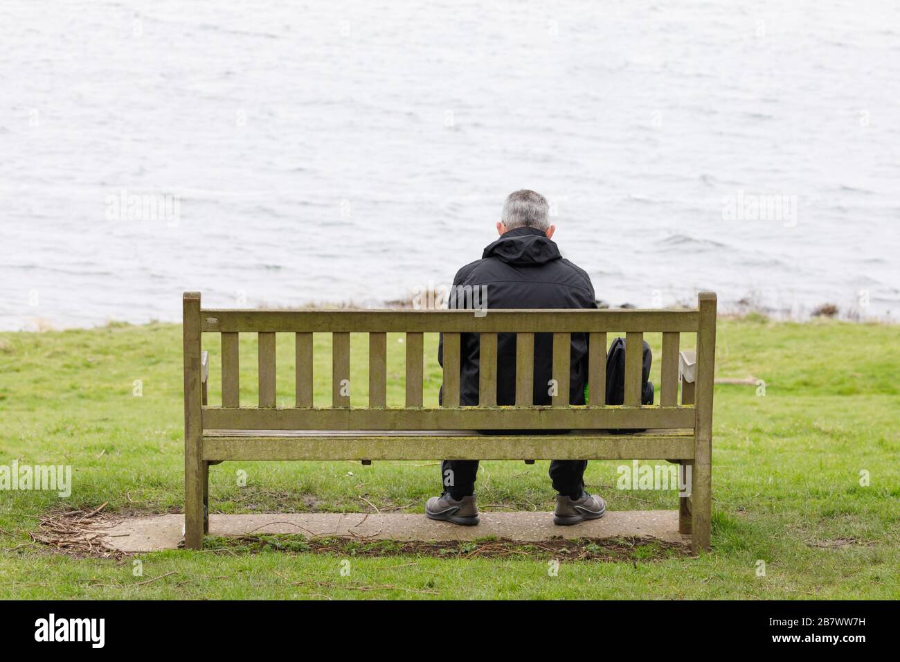 Draycote, Rugby, Warwickshire, March 2020: A man in a waterproof jacket, with his back to us, sits on a wooden bench facing empty water. Stock Photo