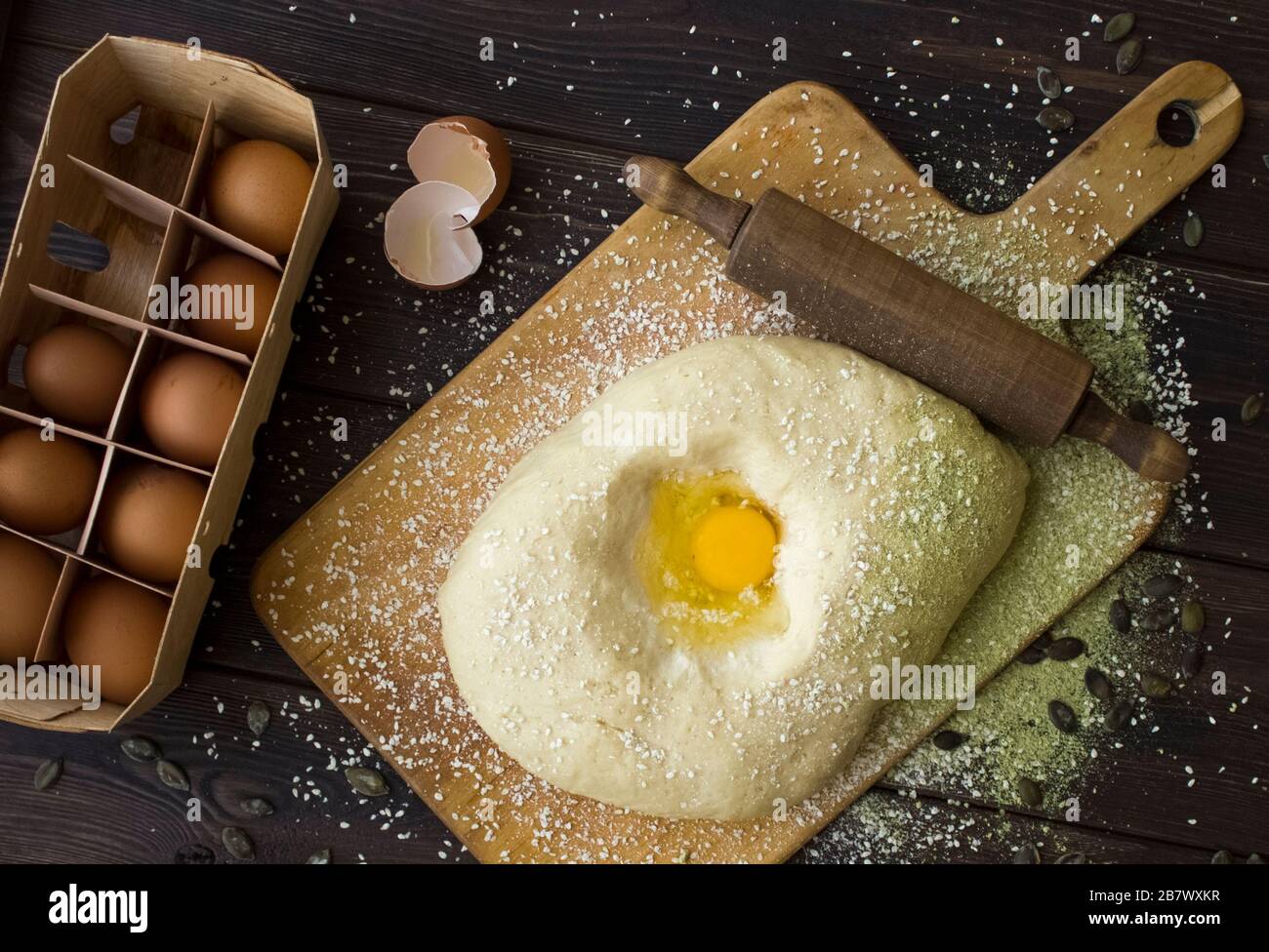 Dough with an egg on a wooden cutting board, eggs and a rolling pin on a dark background. Top view Stock Photo