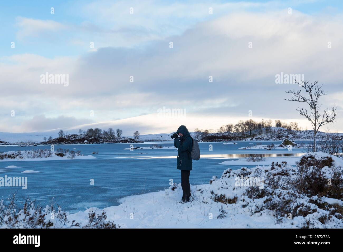 Photographer taking photos on cold winter afternoon with partially frozen loch and snow at Loch Ba, Rannoch Moor, Argyll and Bute, Scotland in January Stock Photo