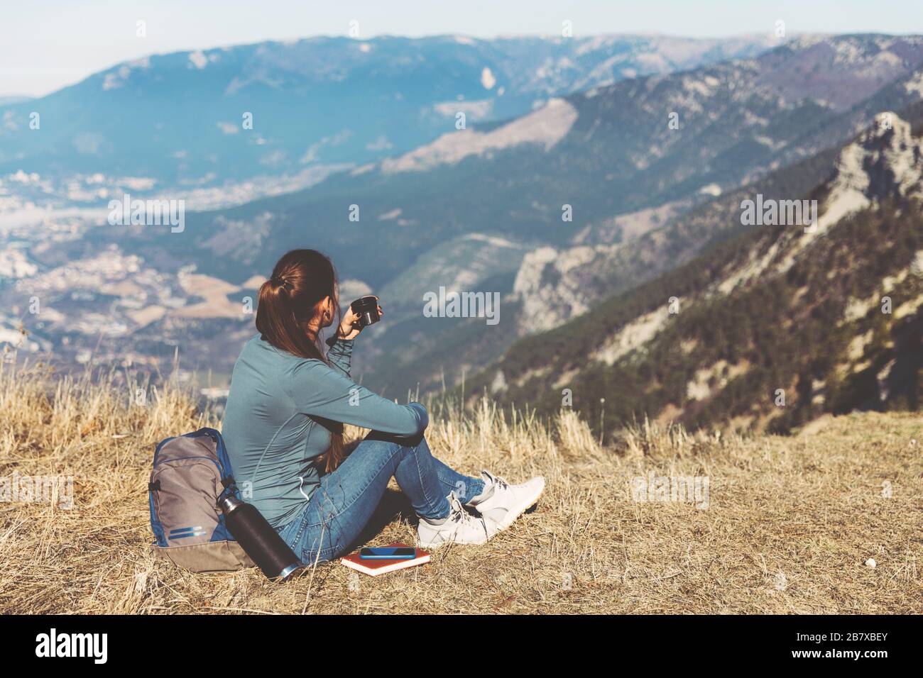 Young beautiful girl travels alone in the mountains in spring or autumn, sits on the edge of the mountain and looks into the distance and enjoys nature, rocks and green forests, view of the landscape. a backpack behind and sportswear, a thermos with a hot drink or tea, freedom and lightness. Back view of woman traveler in cap sitting on mountain top alone and looking at beautiful summer landscape and blue sea view. Drink tea or coffe Stock Photo