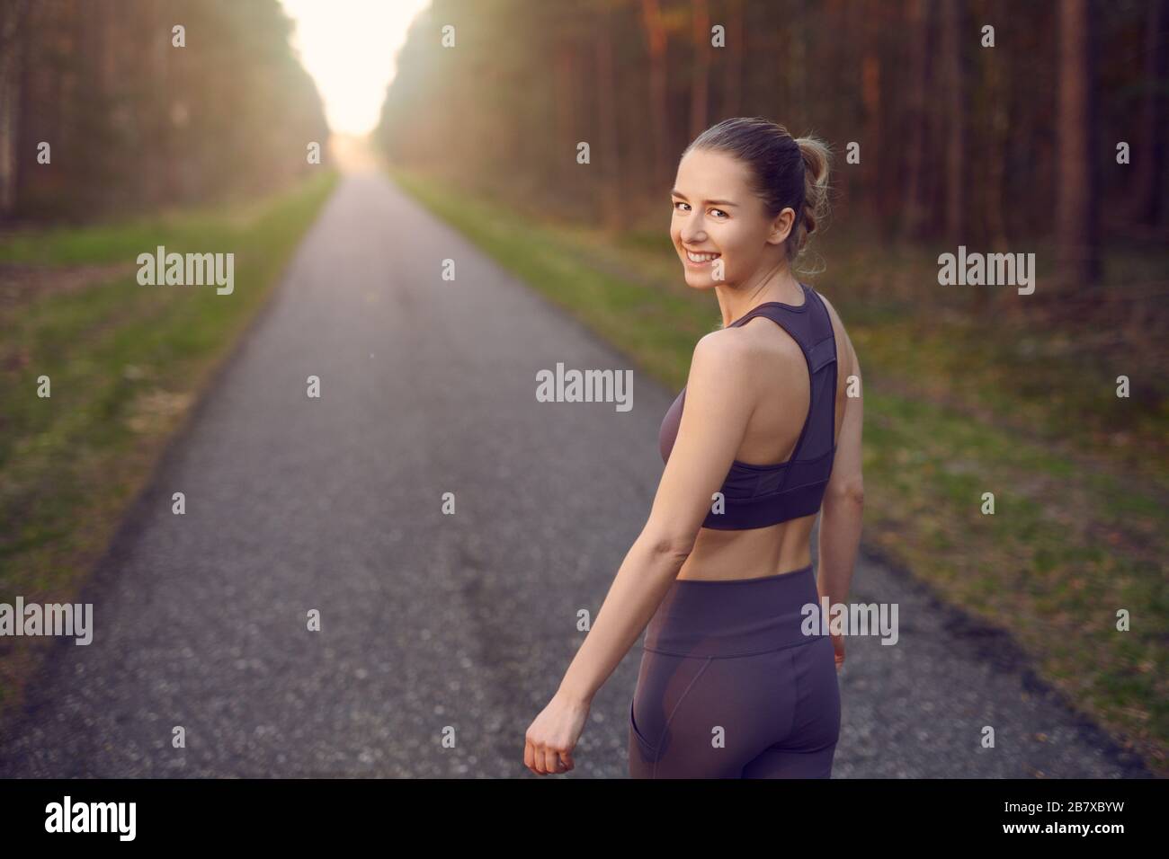 Fit athletic young woman out jogging at sunrise along a straight receding road through forest trees with glow of the sun at the end turning to smile a Stock Photo