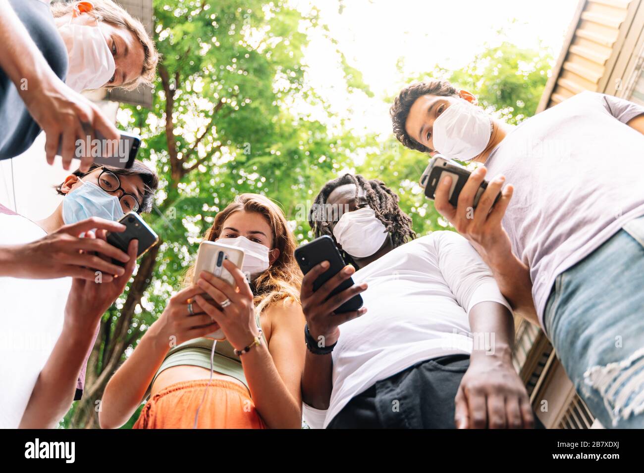 Group of young people of different ethnicities standing together with a mobile phone in hand wearing protective masks on the street Stock Photo