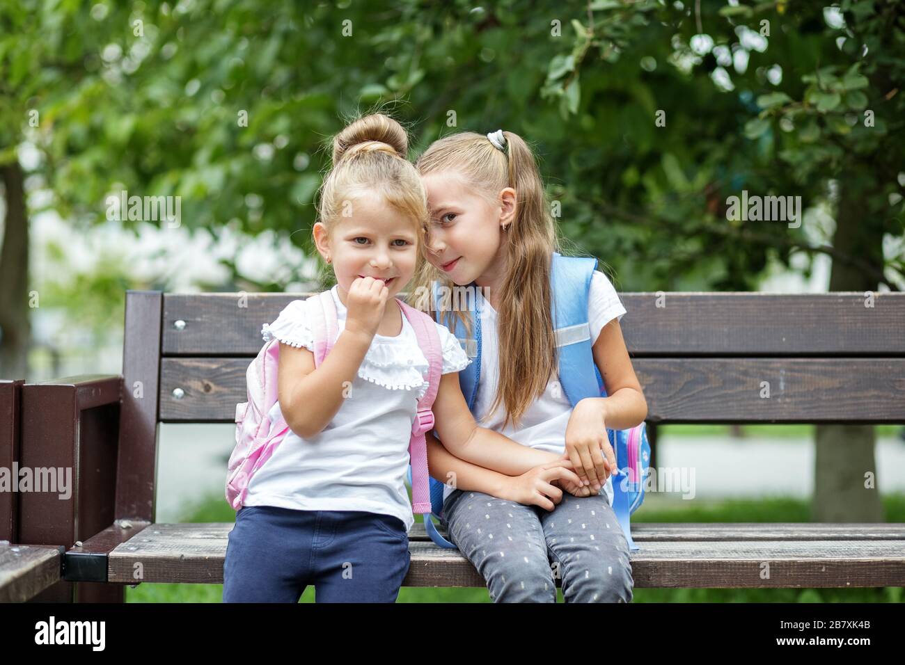 Two small children tell secrets on a bench. Girls with backpacks. The concept is back to school, family, friendship and childhood. Stock Photo