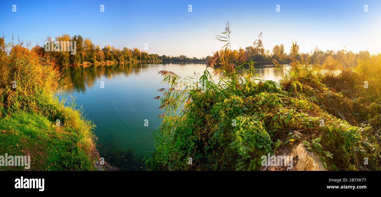 Lake with clear blue water surrounded by trees and vegetation before sunset, panoramic view Stock Photo