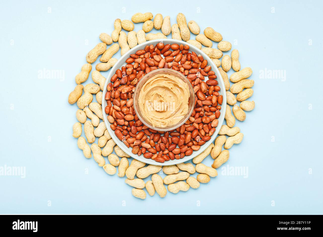 Creamy peanut paste in open jar on peanuts. Near the peanut butter lies peanuts in the shell and peeled peanuts. Minimalistic food flat lay on color Stock Photo