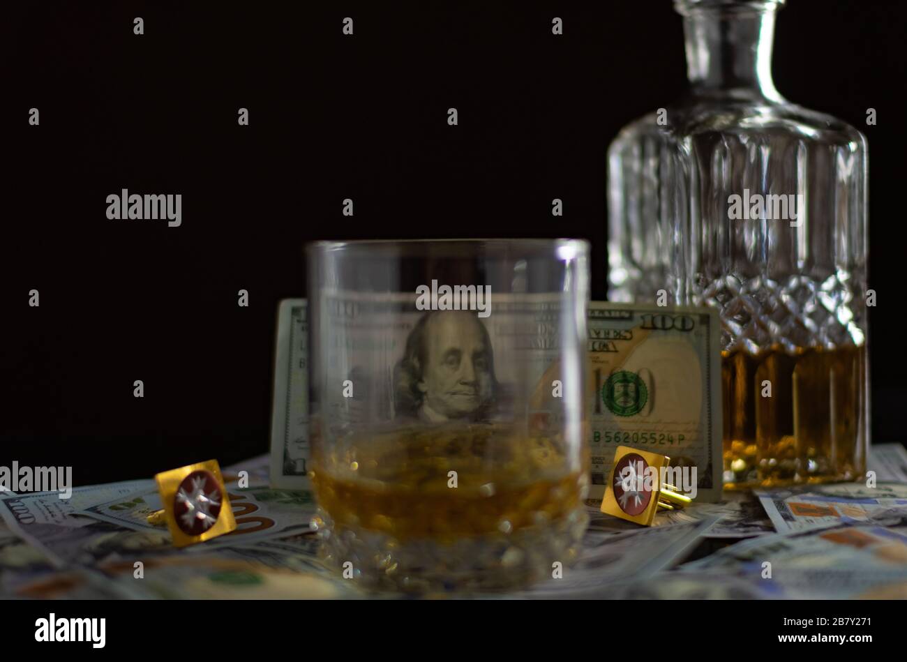 Carafe with whiskey and scattered one hundred dollar bills on the table. Banknote shines through a glass of whiskey Stock Photo