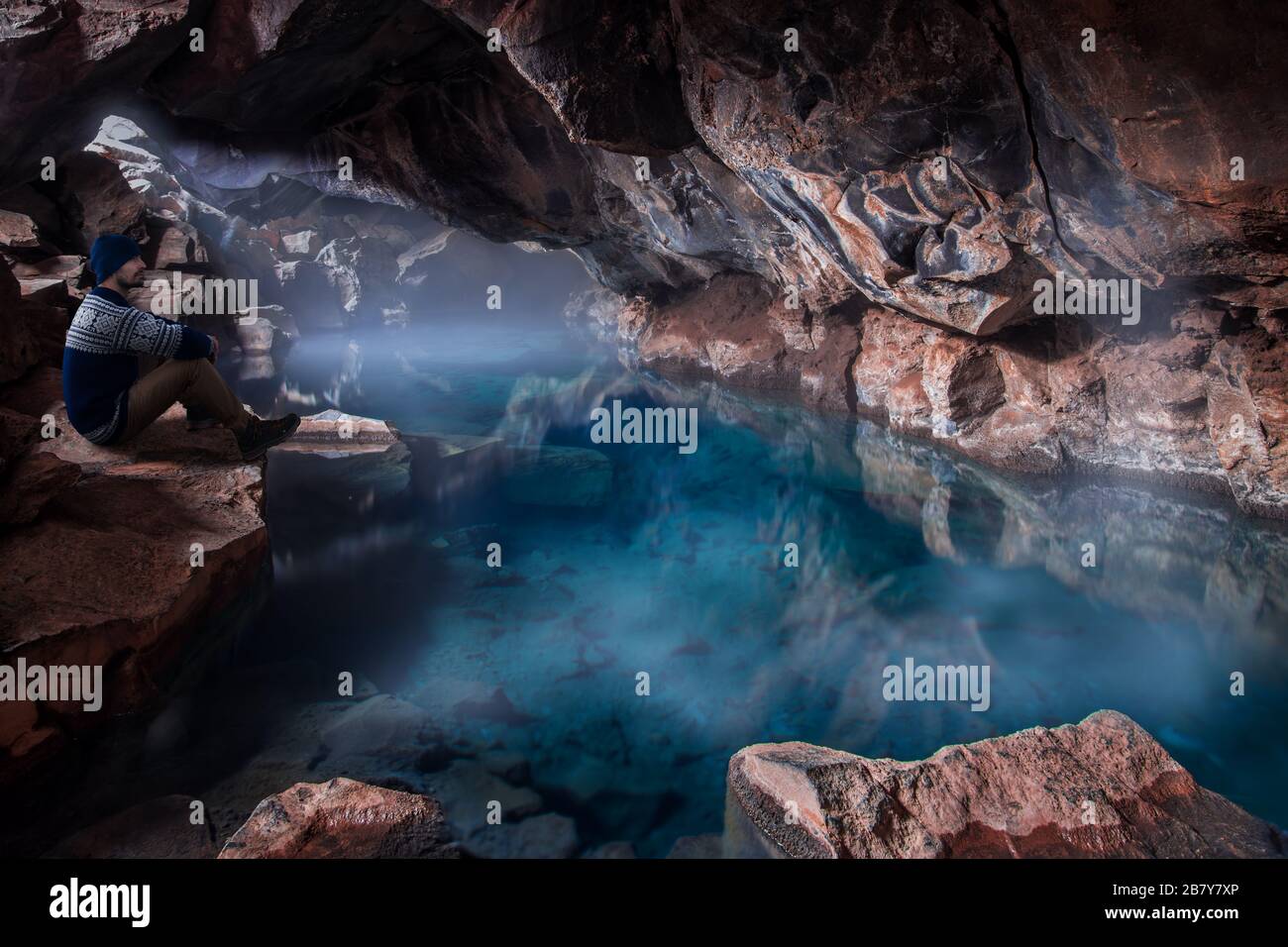 man in traditional Icelandic woolen sweater sitting by beautiful turquoise clear geothermal pool with mist on top inside volcanic cave in Grjotagja, I Stock Photo