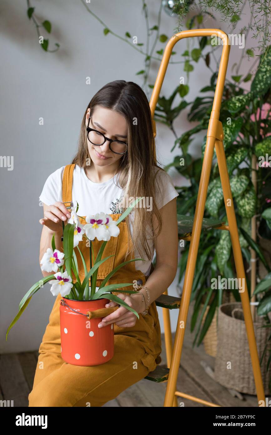 Young smiling woman gardener in glasses wearing overalls, taking care for orchid in old red milk can standing on orange vintage ladder. Home gardening Stock Photo