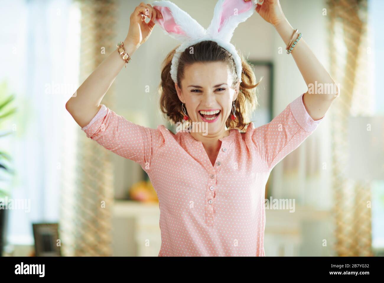 cheerful young housewife in a pink blouse and easter bunny ears in the modern house in sunny spring day jumping. Stock Photo