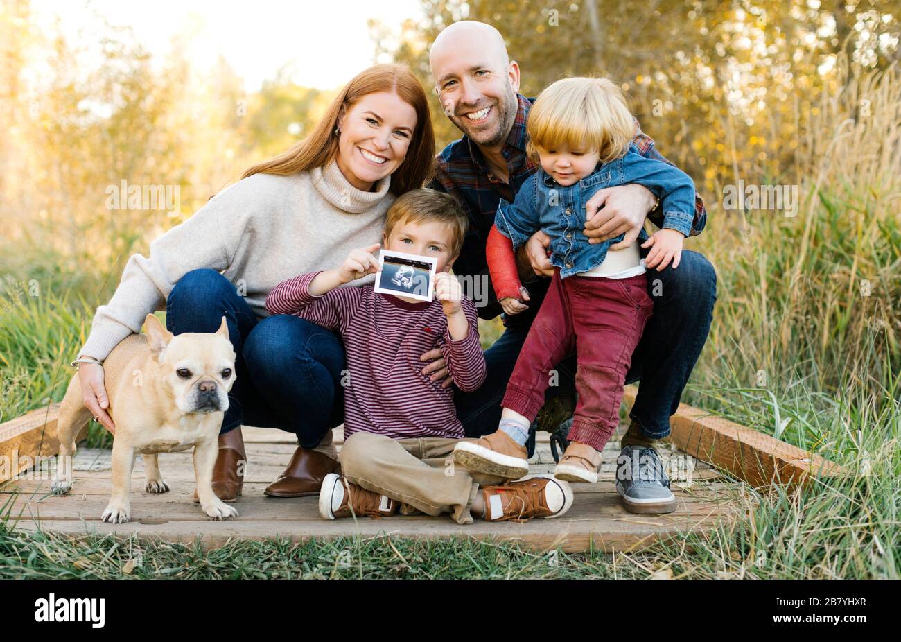 Smiling family with ultrasound photograph and pet dog on forest boardwalk Stock Photo