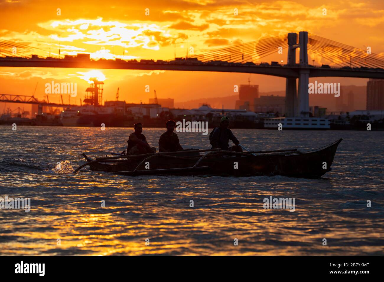 Three men at sunset in a handmade wooden canoe on the Mactan Channel in Cebu with the Marcelo B. Fernan Bridge in the background, Philippines. Stock Photo