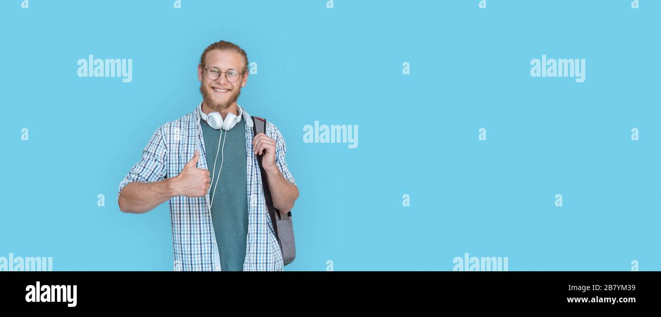 Smiling man student hold backpack thumbs up look at camera isolated on blue. Stock Photo