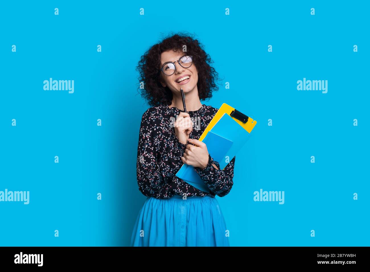 Charming curly haired student holding some books while posing in a dress on blue wall with blank space Stock Photo