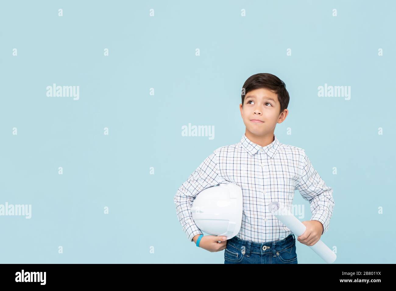 Smiling young asian boy aspiring to be future engineer holding white hardhat and blueprint looking upward in light blue isolated studio backgorund Stock Photo
