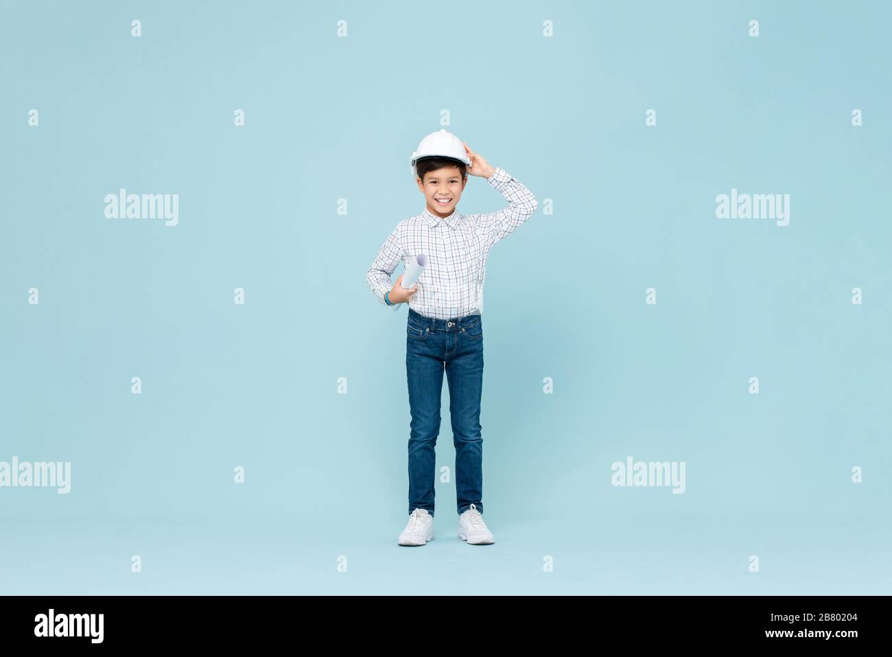 Full length portrait of smiling young asian boy aspiring to be future engineer wearing white hardhat holding blueprint in blue isolated studio backgro Stock Photo