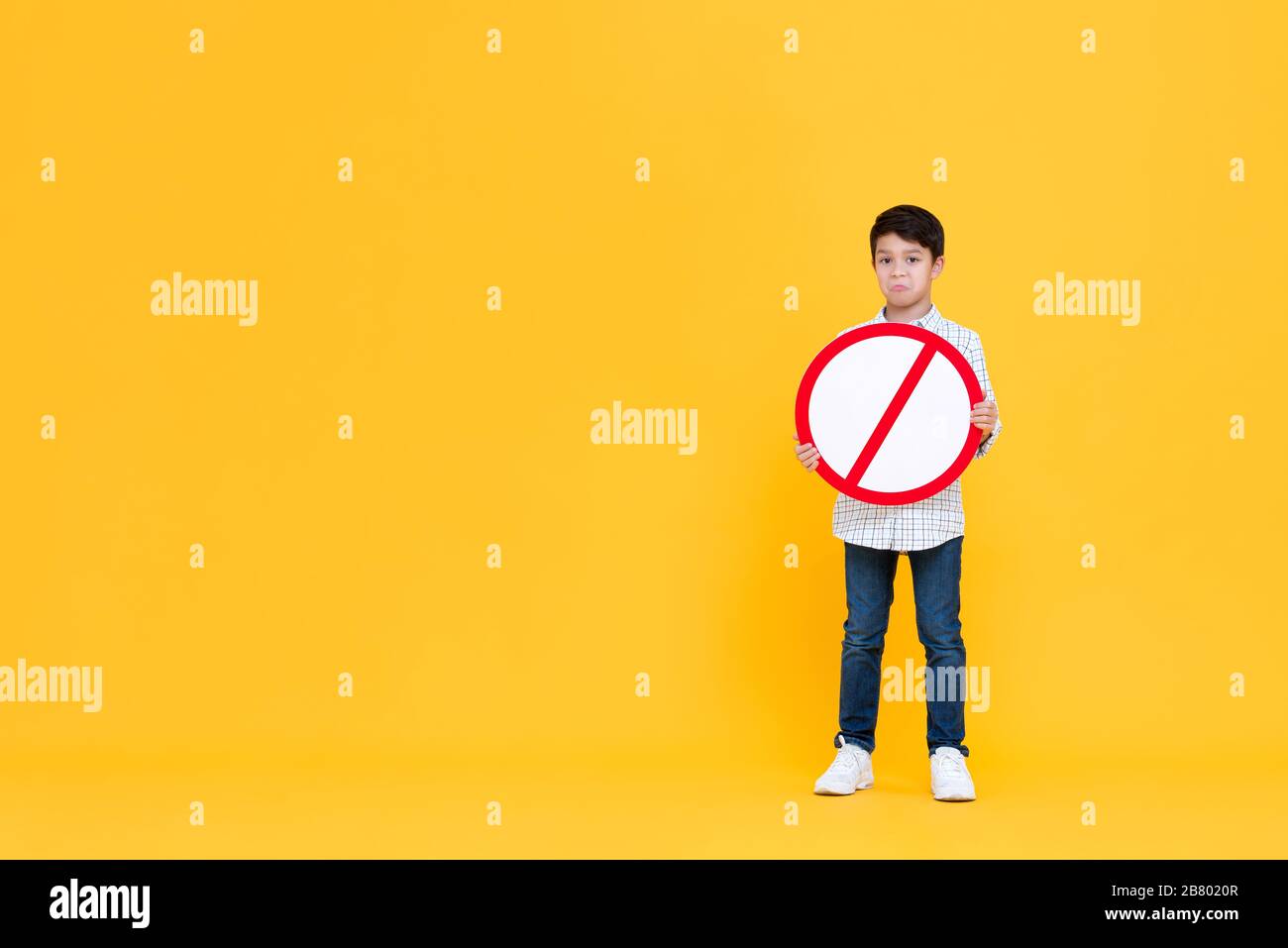 Portait of standing pouting young Asian boy holding red ban signage in yellow isolated studio background with copy space Stock Photo