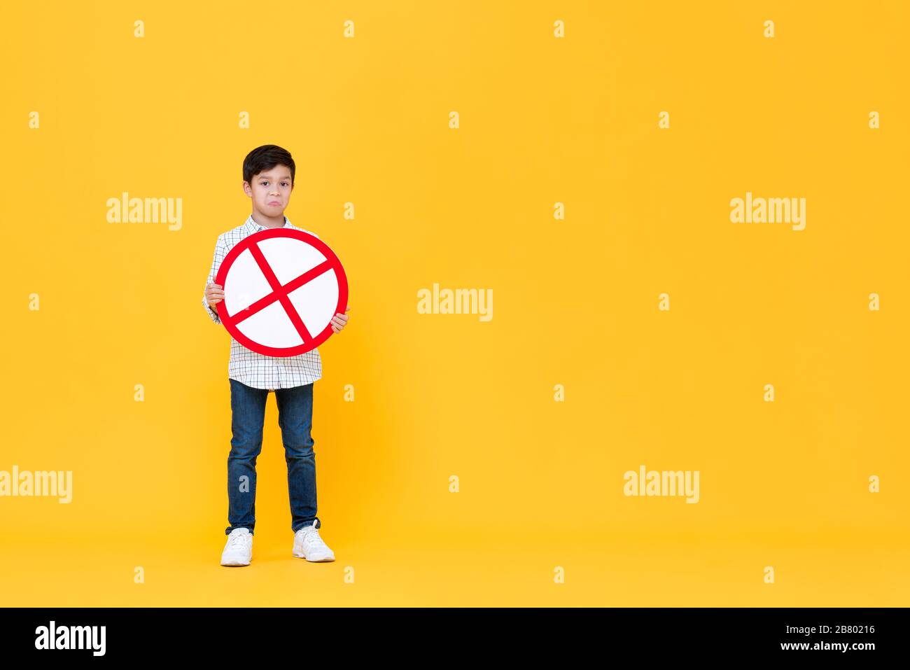 Full length portrait of Asian boy holding red stop or no sign in blank yellow isolated studio background Stock Photo