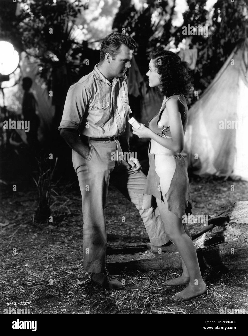 NEIL HAMILTON as Harry Holt and MAUREEN O'SULLIVAN as Jane Parker pose for publicity still with crew member and movie light in background during filming of TARZAN AND HIS MATE 1934 directors CEDRIC GIBBONS and JACK CONWAY characters EDGAR RICE BURROUGHS Photo by TED ALLAN Metro Goldwyn Mayer Stock Photo