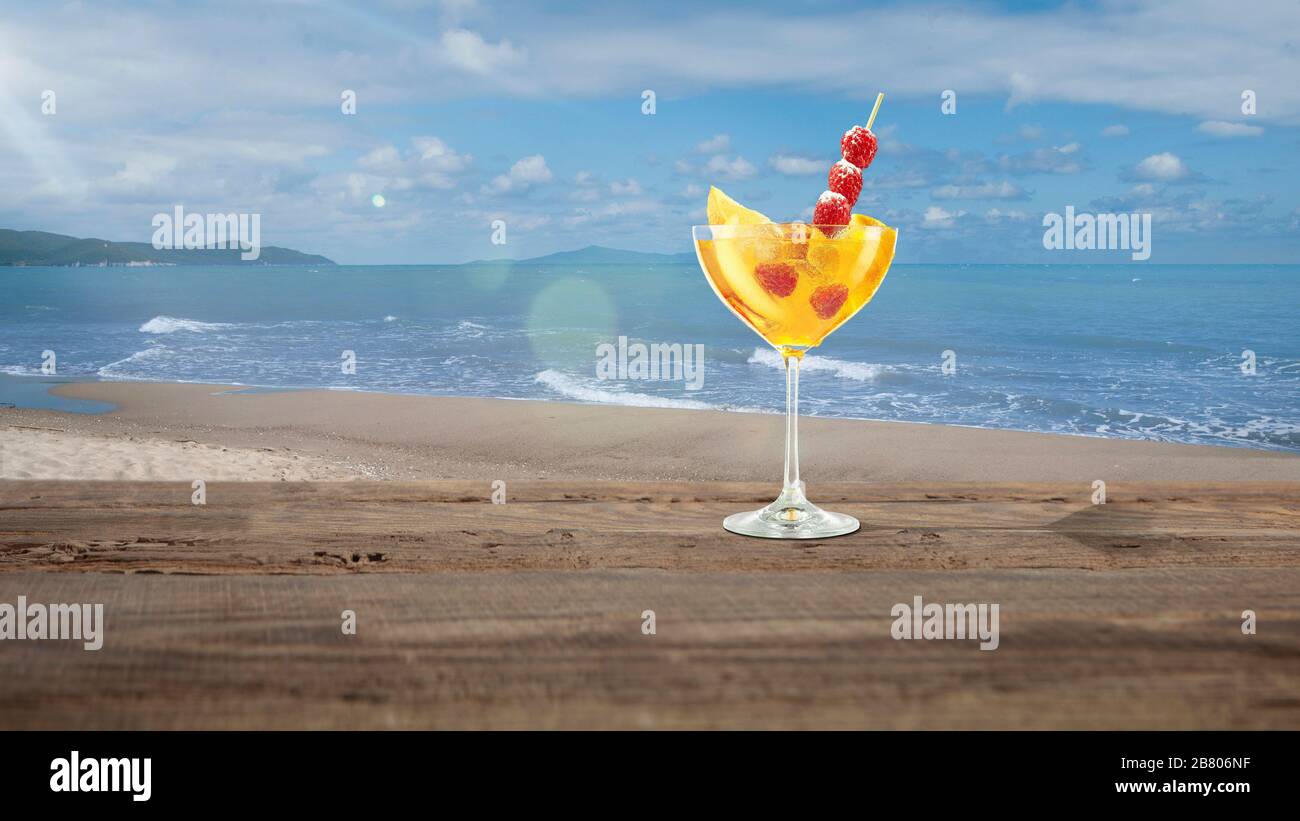 Sunny. Summer bright and tasty refreshing cocktail with sea or ocean beach with palms and white sand on background. Yellow alcohol drink with berries. Concept of summertime, vacation, resort, chill. Stock Photo