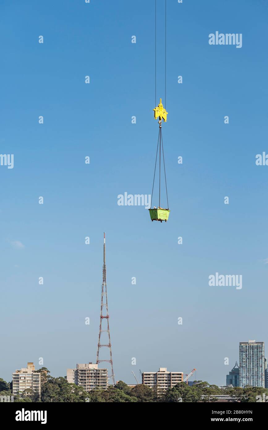 A Skip bin or open-topped waste container is lifted by an overhead tower crane through clear blue sky on a construction site in Australia Stock Photo