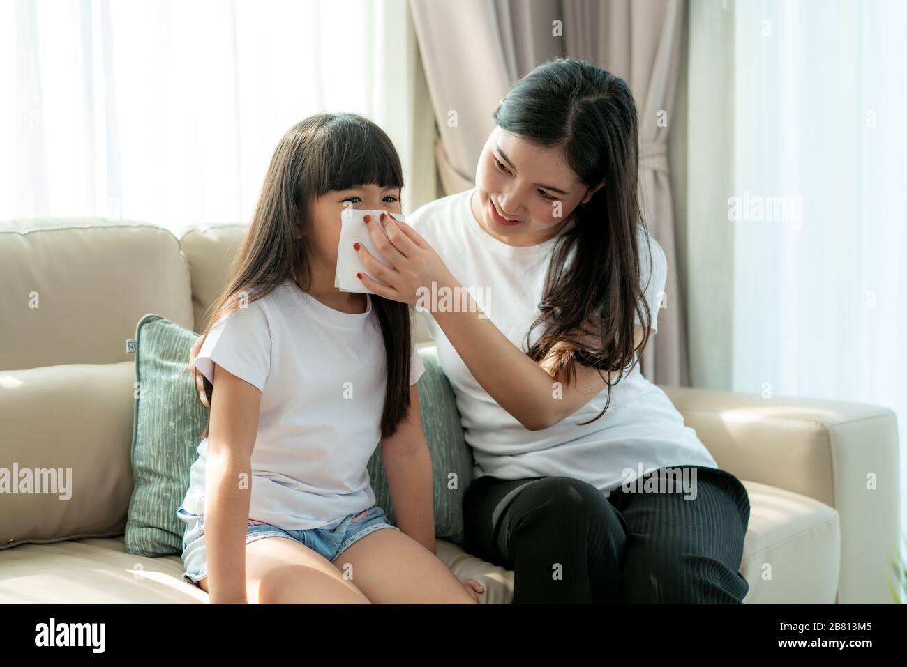 Portrait of cut Asian girl blowing snot into the napkin with her mother is keeping it near her nose with care. Visuals of people feeling sick and suff Stock Photo