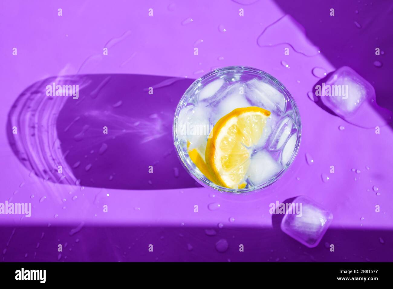 A glass of refreshing water with ice and lemon on a purple background. Heat concept, fresh. Natural light. Flat lay, top view, copy space Stock Photo