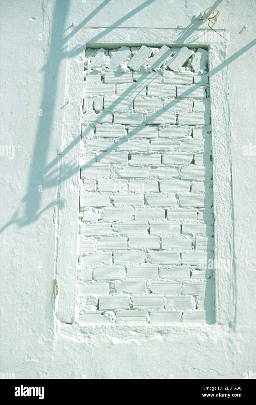 A bricked up window, set high up on the white, stone wall of a building in Parga, Preveza, Epirus, Greece. The electricity wires above cast a shadow on the wall. Stock Photo