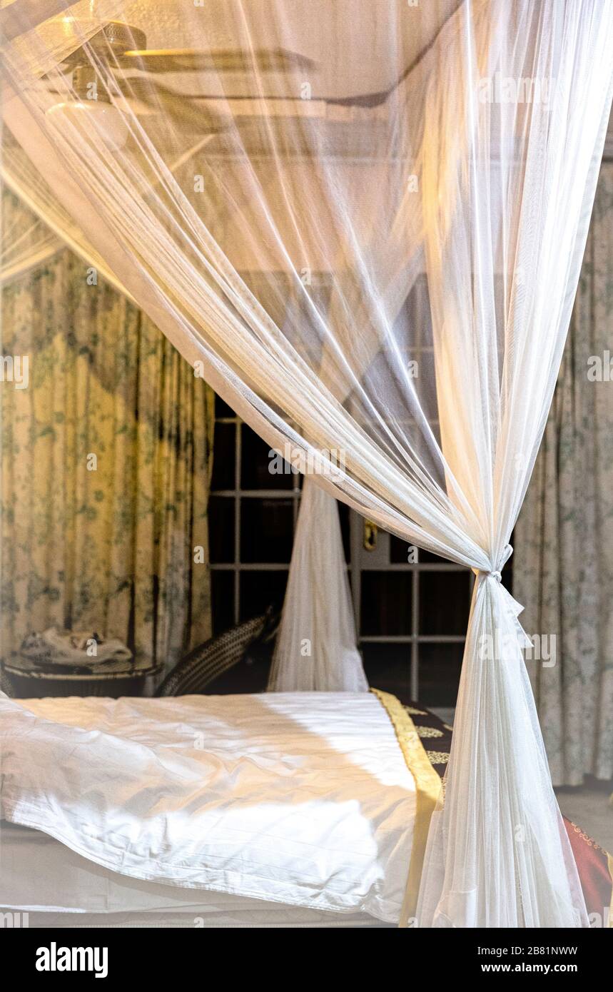 Evocative view across a dimly lit bedroom with detail of the four poster bed, tied mosquito netting and darkened window. Stock Photo